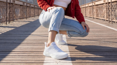Gros plan sur les jambes d'une personne chaussée de baskets Giesswein blanches à semelle texturée, sur une passerelle en bois baignée de soleil. Le logo Giesswein est visible sur le côté de la chaussure.