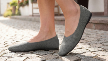 Gros plan sur des pieds chaussés de ballerines en maille grise Giesswein, posés sur un pavé.
