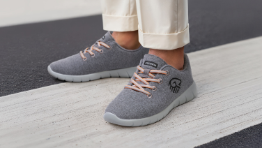 Gros plan sur les pieds d’une personne portant un pantalon crème retroussé. Elle porte des sneakers en laine mérinos grises avec des lacets couleur abricot et une semelle gris clair. La personne se tient sur un passage piéton.
