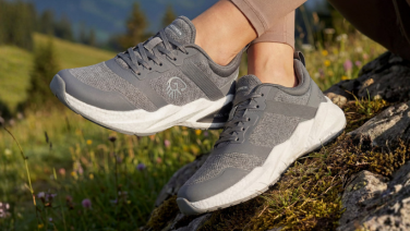 Gros plan sur les pieds d'une personne debout sur un rocher recouvert de mousse, dans un paysage de montagne. Elle porte des baskets grises de sport avec une semelle extérieure blanche.