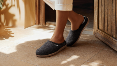 Des pieds chaussés d'un pantalon blanc et de chaussons en laine gris foncé posés sur un carrelage clair, la lumière chaude du soleil illumine le sol.