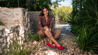 Une femme en tailleur marron est assise devant un mur de pierre dans un jardin méditerranéen ; elle porte des ballerines rouges Giesswein.