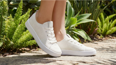 Gros plan sur des pieds chaussés de baskets en tricot Giesswein blanches à semelle blanche, posés sur un chemin de galets au milieu de fougères vertes.