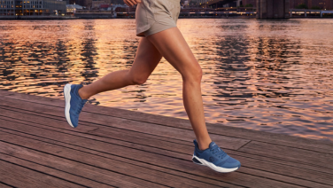 Une personne vêtue d'une tenue de sport beige court sur une passerelle en bois au bord de l'eau au coucher du soleil ; on distingue les contours d'une ville en arrière-plan. Elle porte des chaussures de course bleues et sportives.