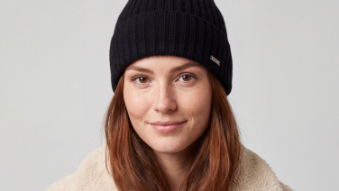 Portrait d'une femme souriante aux cheveux roux, coiffée d'un bonnet noir en tricot côtelé orné d'une étiquette métallique ; elle porte une veste en polaire couleur crème sur un fond gris clair.