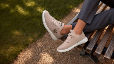 Gros plan sur des sneakers Giesswein en mérinos beige avec semelle blanche et marquage du logo, aux pieds d’une personne en pantalon gris assise sur un banc de parc.