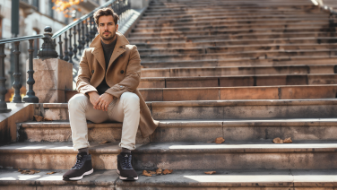 Un homme vêtu d'une veste beige et d'un pantalon clair est assis sur un escalier extérieur en pierre et porte des baskets d'hiver Giesswein noires, ambiance automnale.