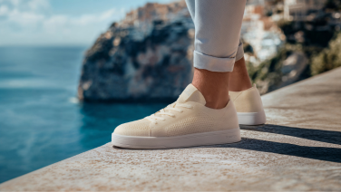 Gros plan d'une sneaker Giesswein en mérinos couleur crème avec semelle plate blanche sur un muret de pierre, devant un décor marin méditerranéen.