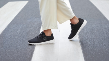 Gros plan sur des sneakers Giesswein en mérinos noirs avec semelle blanche aux pieds d’une personne portant un pantalon large blanc, sur un passage piéton.