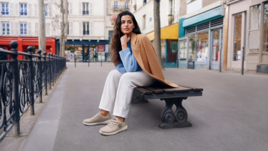 Une femme est assise sur un banc public dans le centre-ville, vêtue d'une veste beige, d'un t-shirt bleu, d'un pantalon blanc et de baskets Giesswein Merino Runners beiges.