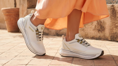 Gros plan sur les jambes d'une femme vêtue d'une jupe orange, debout sur un pavé baigné de soleil, chaussée de baskets blanches Giesswein ornées de détails dorés et dotées d'une semelle texturée.