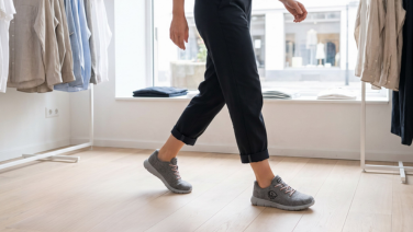Une personne vêtue d'un pantalon foncé aux ourlets retroussés traverse une boutique lumineuse au sol en parquet ; à l'arrière-plan, des vêtements sont suspendus à des portants. Elle porte des baskets grises en laine mérinos à lacets.