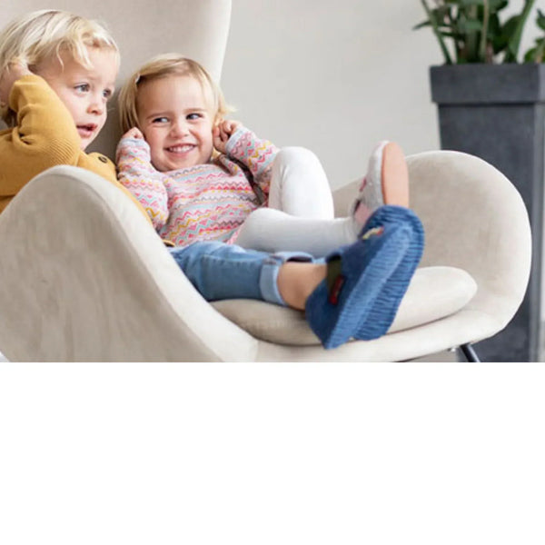 Deux jeunes enfants sont assis ensemble dans un fauteuil beige, l'enfant à l'avant porte des chaussons tricotés bleus et sourit joyeusement.