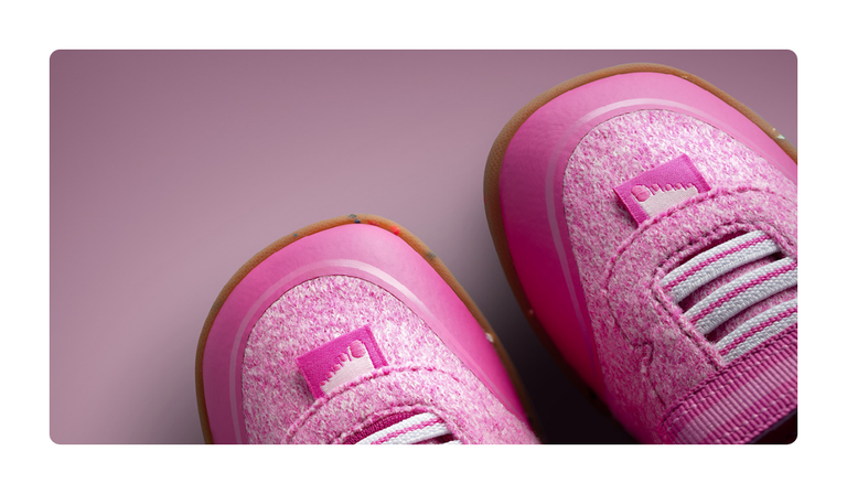 Gros plan sur deux chaussures pour enfants Giesswein de couleur rose, avec une tige en laine mérinos, des lacets élastiques et une semelle couleur miel, sur fond rose.