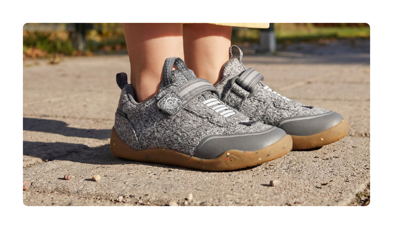 Gros plan sur des baskets pour enfants Giesswein grises, avec une tige en laine mérinos, une fermeture velcro et une semelle couleur miel, sur un chemin de sable.